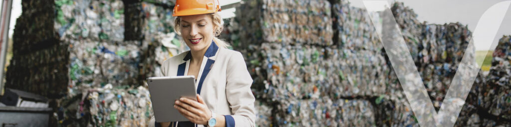 Una mujer ingeniera inspecciona datos en una tablet frente a grandes pilas de materiales reciclables compactados, probablemente botellas de plástico. La escena captura la integración de la tecnología en el reciclaje de recursos y el compromiso con prácticas sostenibles.